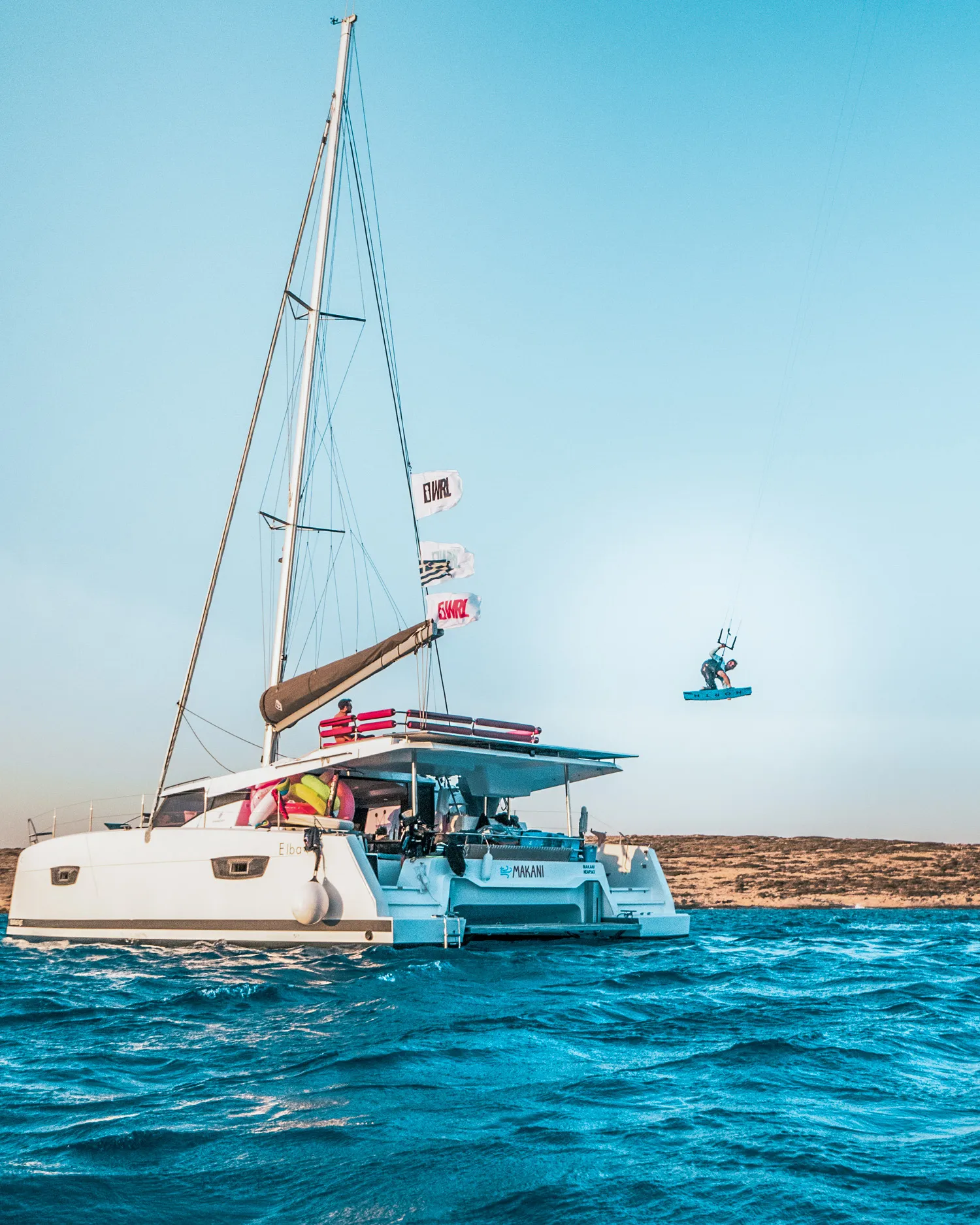 A kitesurfer jumping high beside Makani at anchor, the boat flying its flags in the late sun.