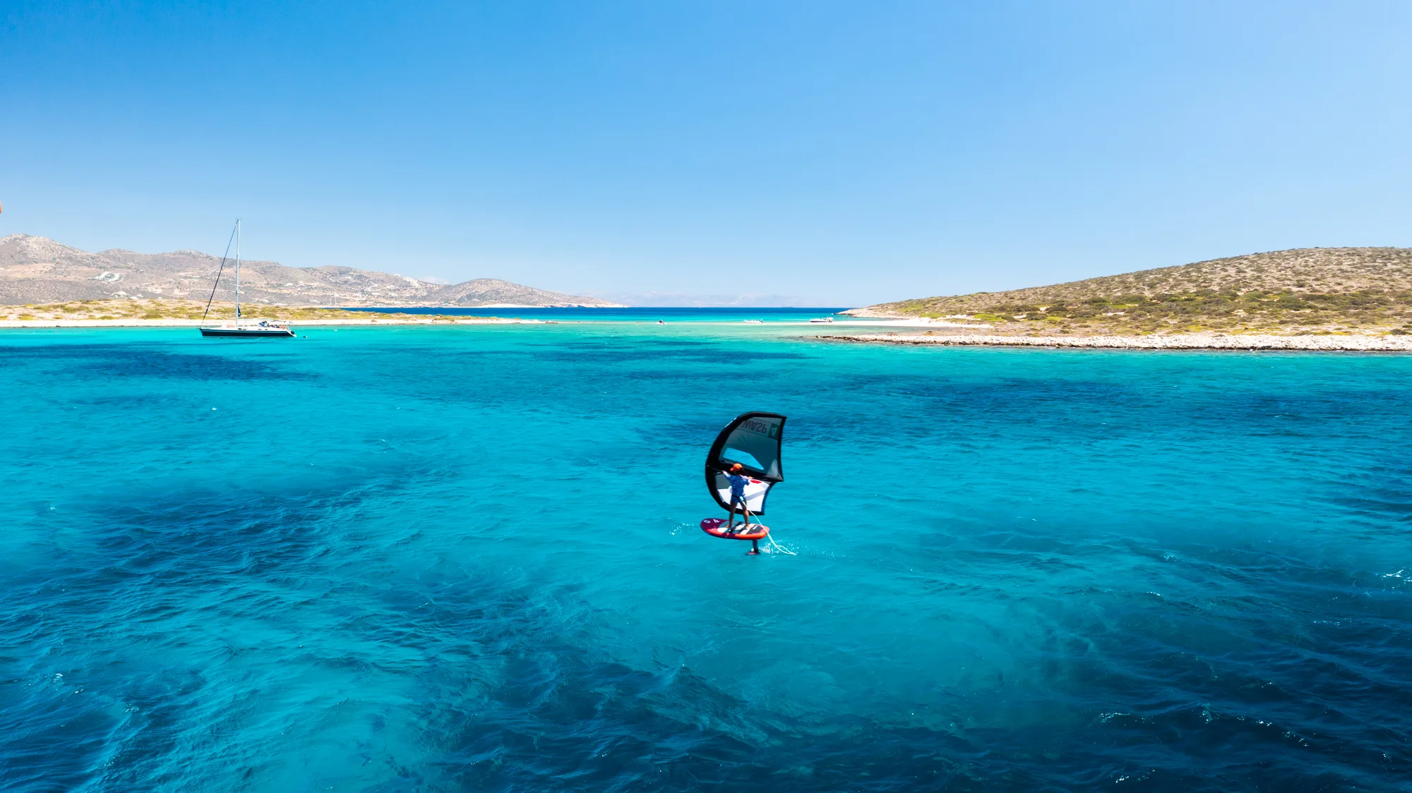Wing foiler on crystal-clear turquoise water with a sailboat and Cyclades islands in the background.